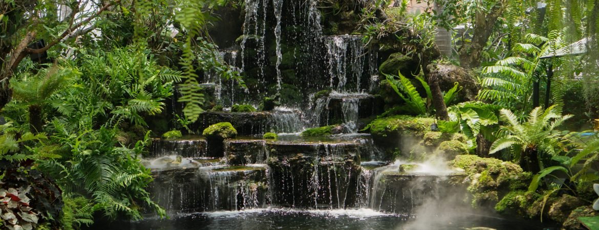View of a beautiful artificial waterfall surrounded by lush green tropical trees.