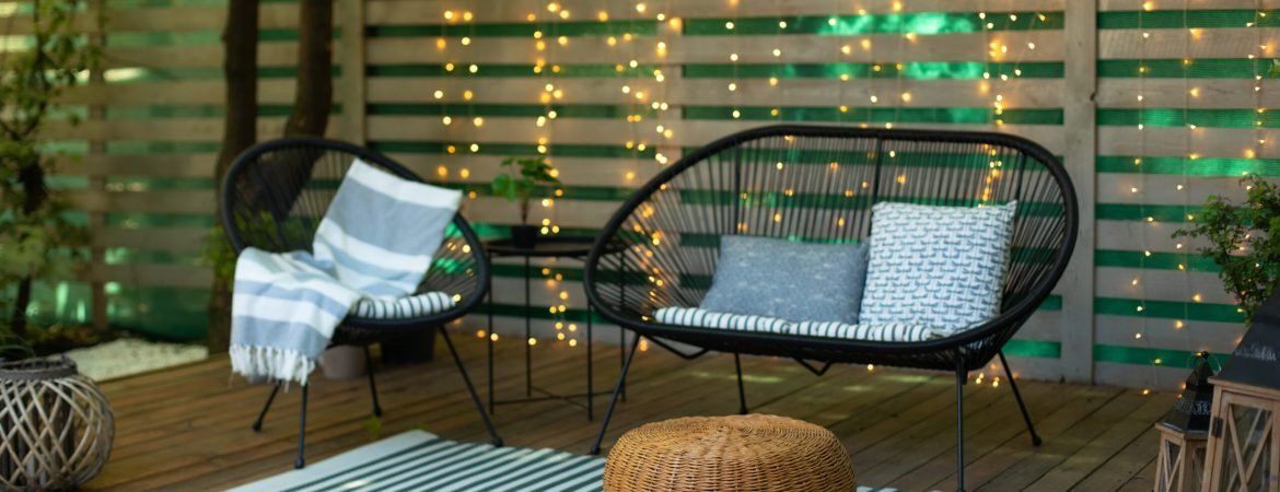 Terrace house with plants, wooden wall and table, comfortable sofa, armchair and lanterns. Cozy space in patio or balcony. Wooden veranda with garden furniture. Modern lounge outdoors in backyard