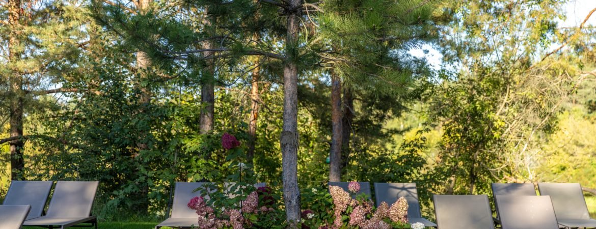 This image captures a serene outdoor lounging area with gray chairs set against lush trees and vibrant flowering plants, perfect for relaxation and enjoying nature.